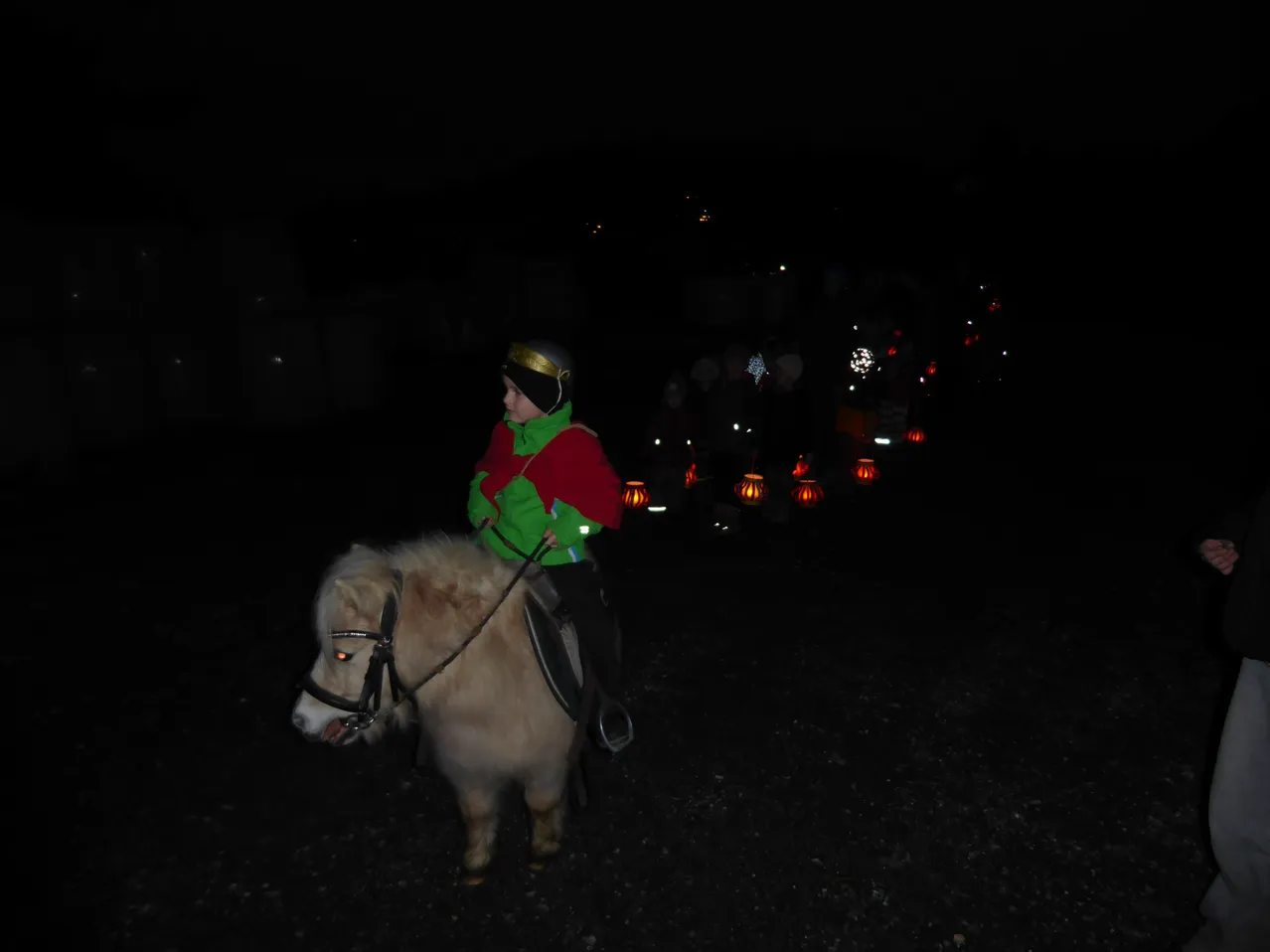 Ein Kind reitet ein kleines Pferd in der Dunkelheit. Sie tragen eine grün-rote Jacke. Hinter ihnen halten Menschen leuchtende Laternen.