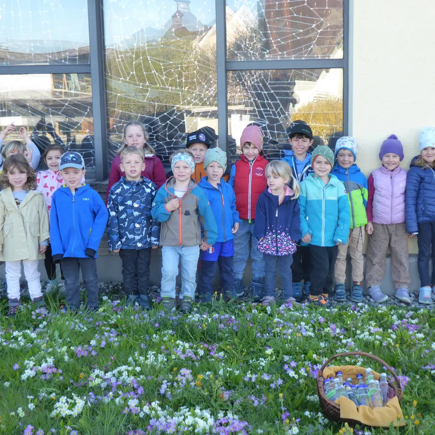 Eine Gruppe von Kindern, in Winterkleidung, steht draußen in einem Blumenfeld mit einem zerbrochenen Glasfenster dahinter.