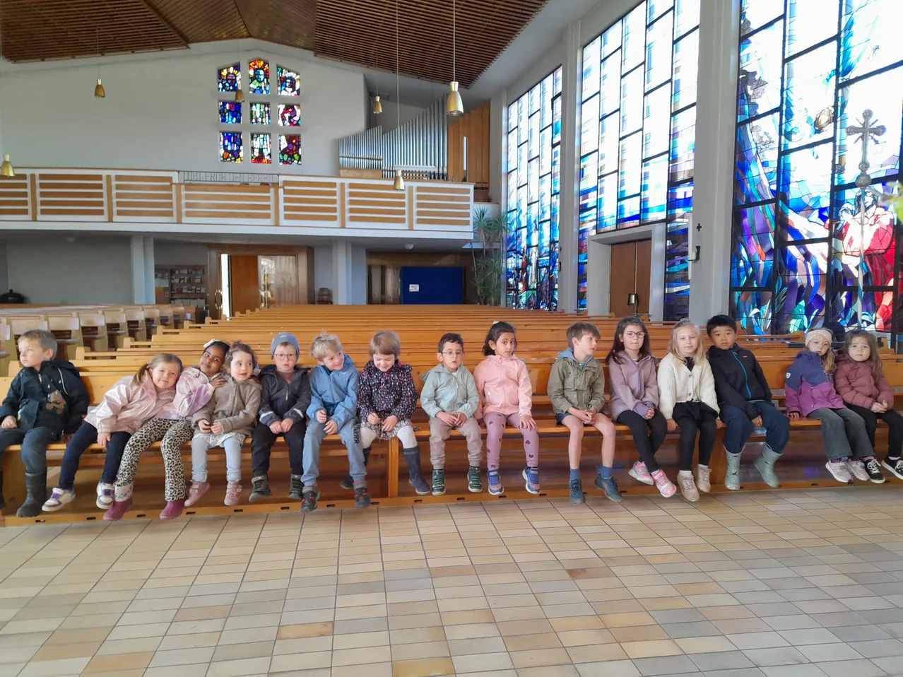 Eine Gruppe von Kindern sitzt auf einer Holzbank in einer Kirche. Die Kirche hat große Buntglasfenster und eine Holzempore.