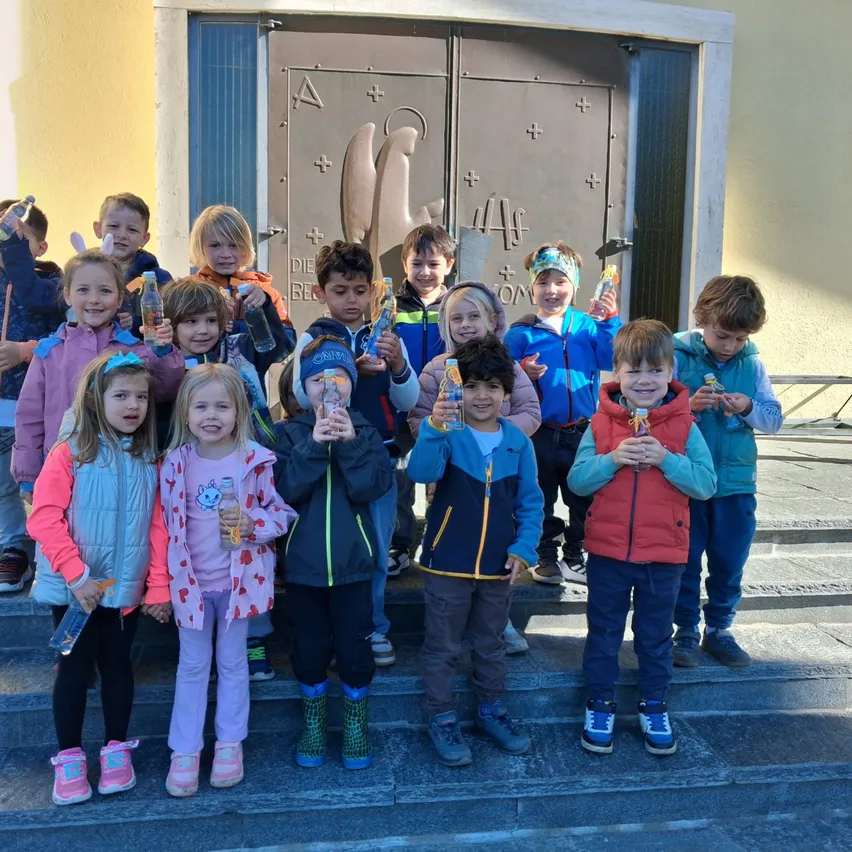 Eine Gruppe von Kindern in Winterkleidung steht auf Stufen und hält Flaschen in der Hand. Dahinter ist ein Gebäude mit einer großen Tür und einem Schild mit dem Buchstaben 'A' zu sehen.