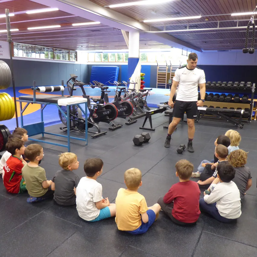 Ein Mann in einem Fitnessstudio demonstriert Übungen für eine Gruppe junger Jungen, die im Kreis sitzen. Hanteln und Trainingsgeräte umgeben sie.