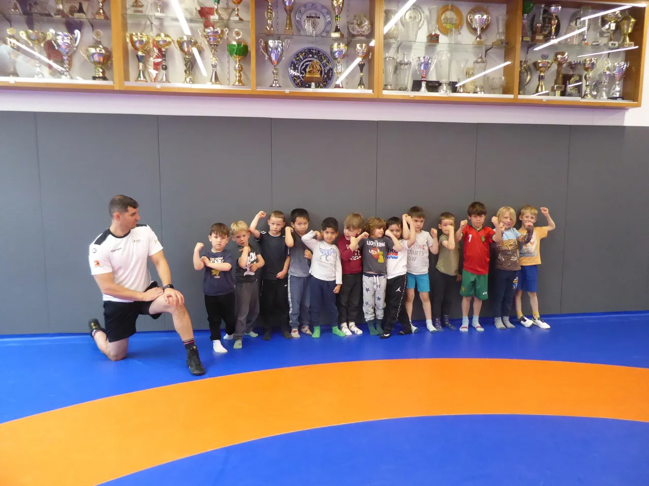Eine Gruppe von Kindern steht in einer Reihe in einer Wrestling-Halle und posiert mit einem Trainer vor einer Wand mit Trophäen-Ausstellungen.