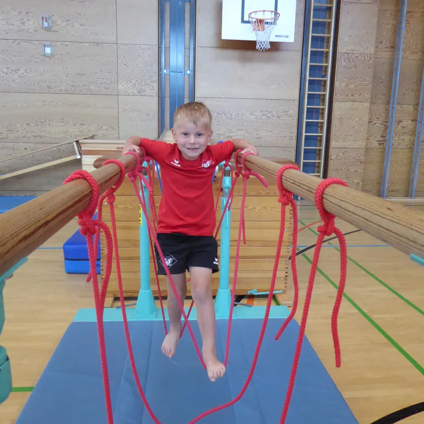 Ein junger Junge hängt an einem Set von Gymnastikringen und trägt ein rotes T-Shirt und schwarze Shorts. Der Fitnessraum hat Holzböden, blaue Matten und einen Basketballkorb im Hintergrund.