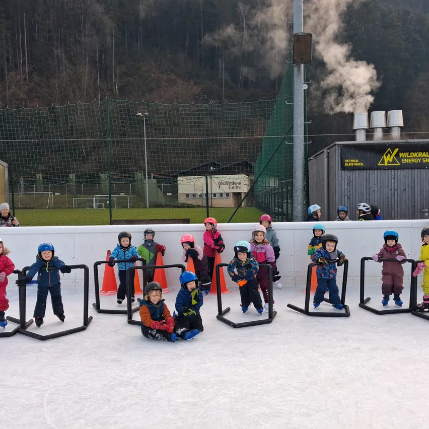Eine Gruppe von Kindern in Winterkleidung und Helmen steht auf einer Eisbahn. Sie halten sich an schwarzen Barrieren fest, mit orangefarbenen Kegeln dahinter. Ein Gebäude mit einer grünen Plane ist im Hintergrund zu sehen.