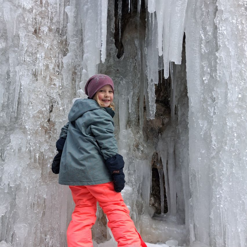 Ein kleines Mädchen in einer Winterjacke, orangefarbenen Hosen und einer lila Mütze steht vor einer großen Eisformation, möglicherweise in einer Eishöhle. Sie lächelt und scheint die winterliche Atmosphäre zu genießen.