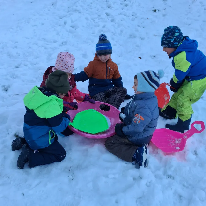 Fünf Kinder spielen im Schnee. Sie tragen Winterkleidung und Mützen. Zwei von ihnen halten Schneeschaufeln, und einer hält eine pinke Schneeschaufel. Sie sitzen im Schnee, und einer von ihnen kniet.