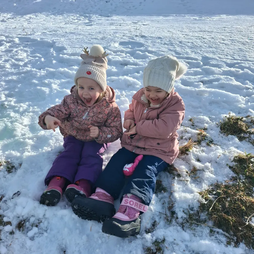 Zwei Kinder sitzen auf einer verschneiten Oberfläche, beide in Winterkleidung und lächeln. Eines hat einen Rentierhut und das andere hat ein rosa Band an den Stiefeln.