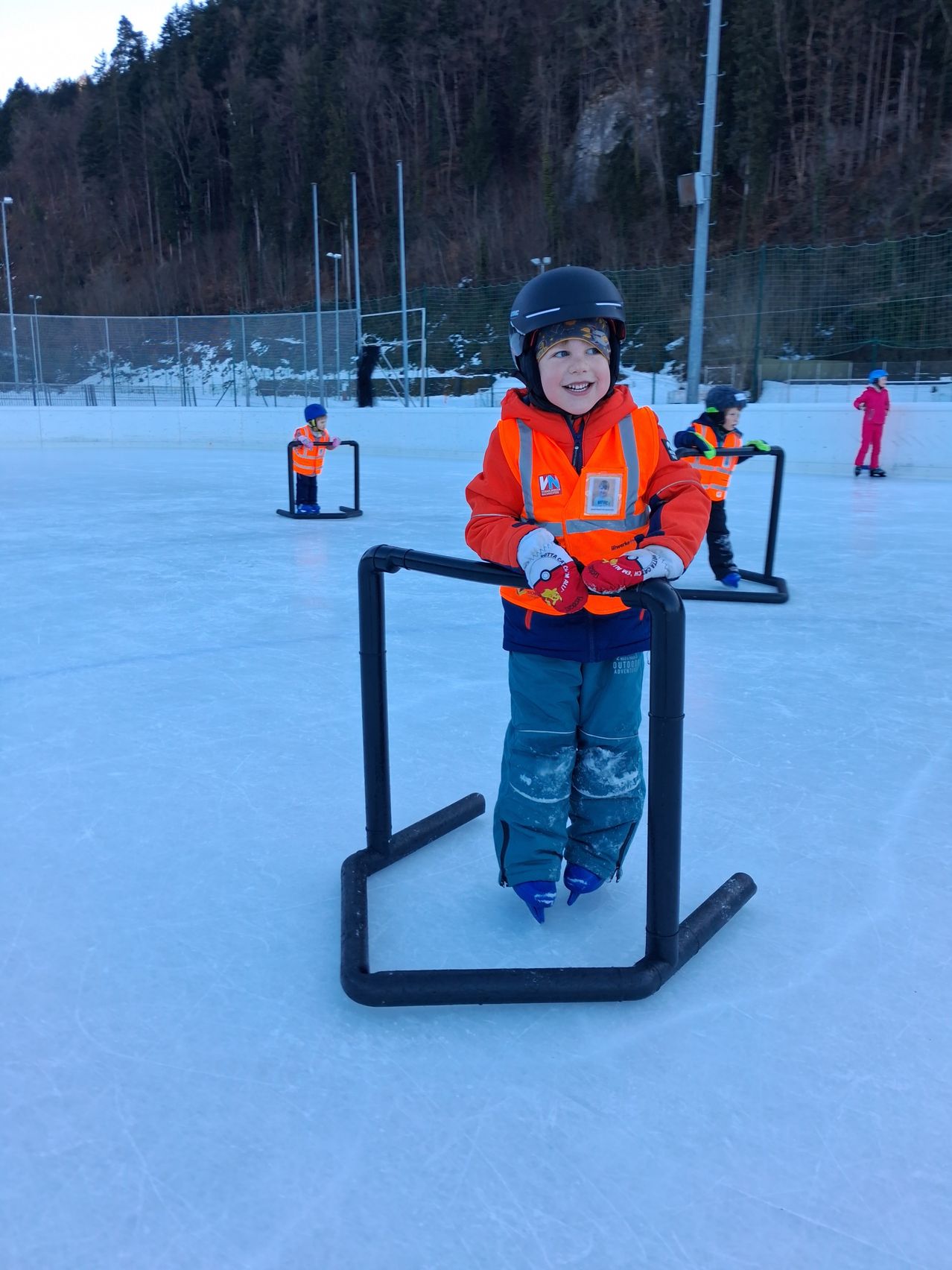 Ein Kind steht in einer Eishalle und hält sich an einem schwarzen Stützbügel fest. Es trägt einen leuchtend orangefarbenen Mantel, einen Helm und Handschuhe. Im Hintergrund sind auch andere Kinder in Winterkleidung beim Eislaufen zu sehen.