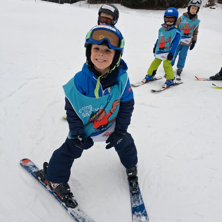 Ein junger Junge steht auf Skiern und lächelt. Hinter ihm sind drei Kinder beim Skifahren, zwei von ihnen tragen blaue Westen. Sie befinden sich auf einem verschneiten Hang.