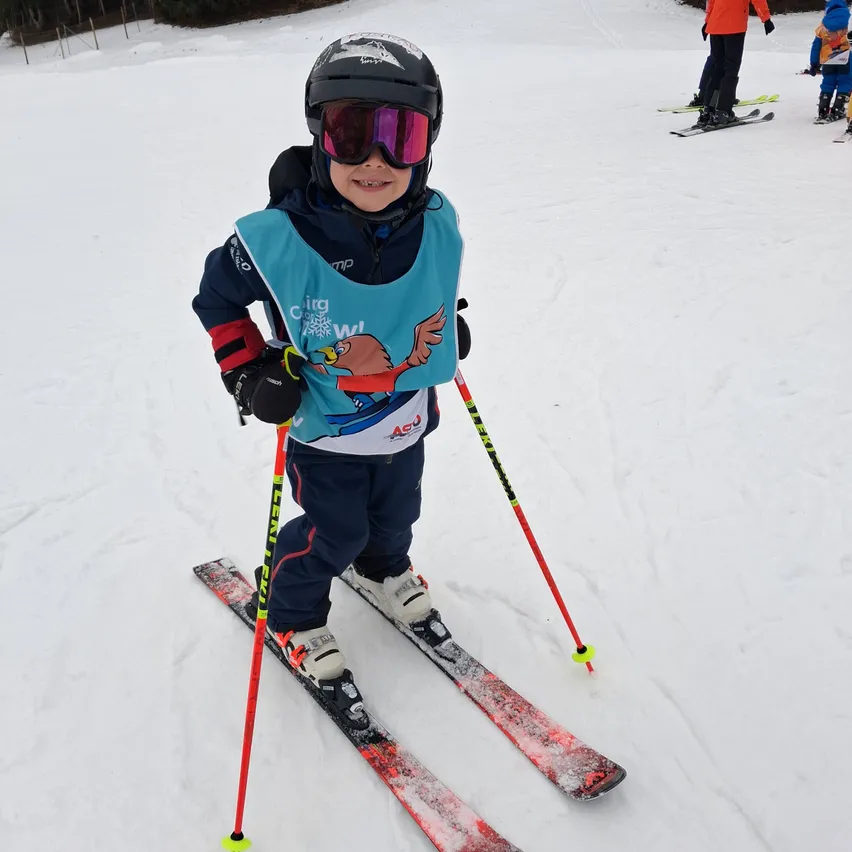 Ein junger Junge, der auf einem verschneiten Berg mit Skiausrüstung und Helm skifahrert. Er trägt eine blaue Weste mit einem Comic-Charakter und hält rote Skistöcke.