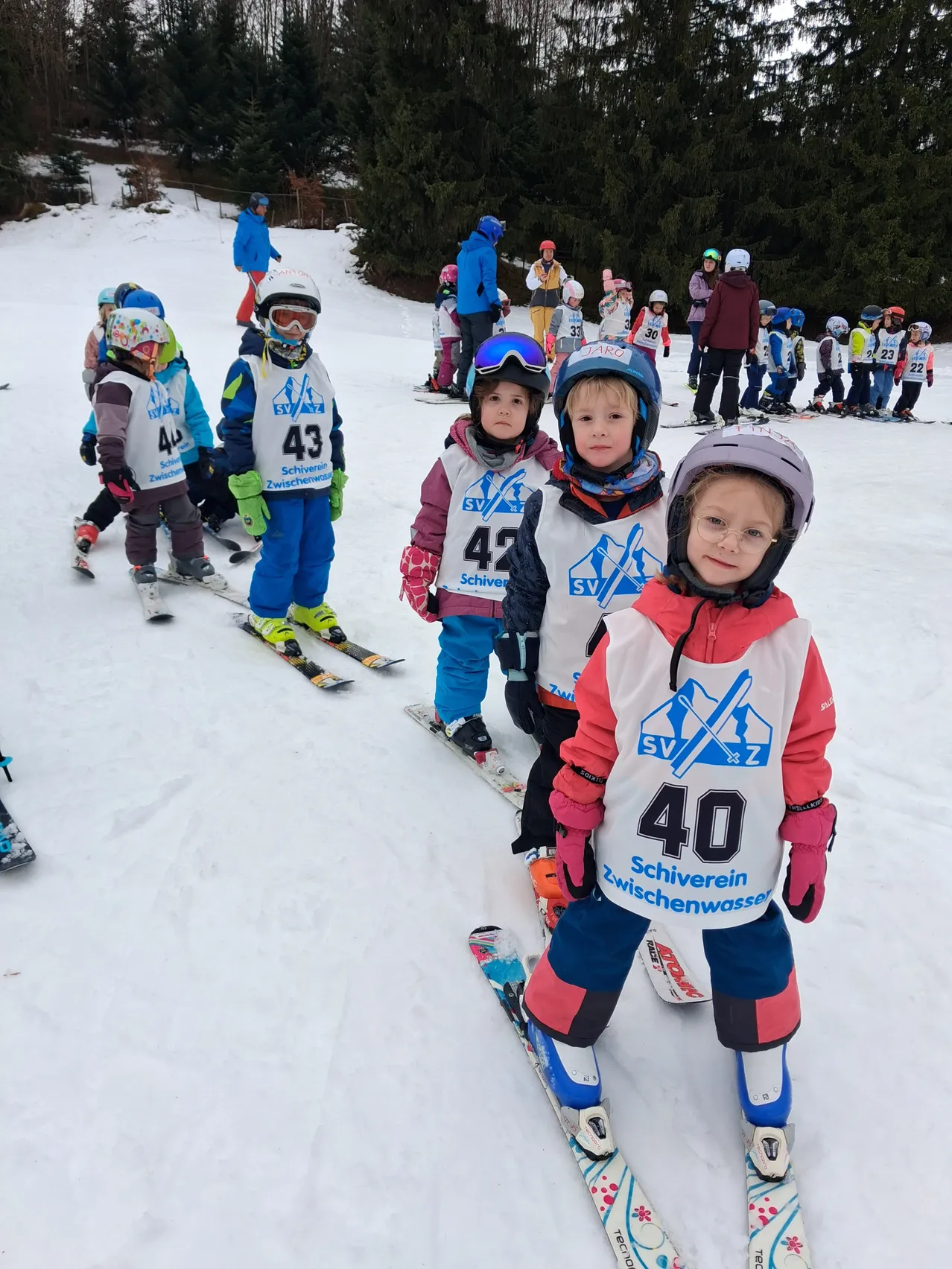Eine Gruppe von Kindern in Skiausrüstung, mit den Nummern 40 bis 43, steht in einem verschneiten Bereich. Sie tragen Skijacken und Helme.