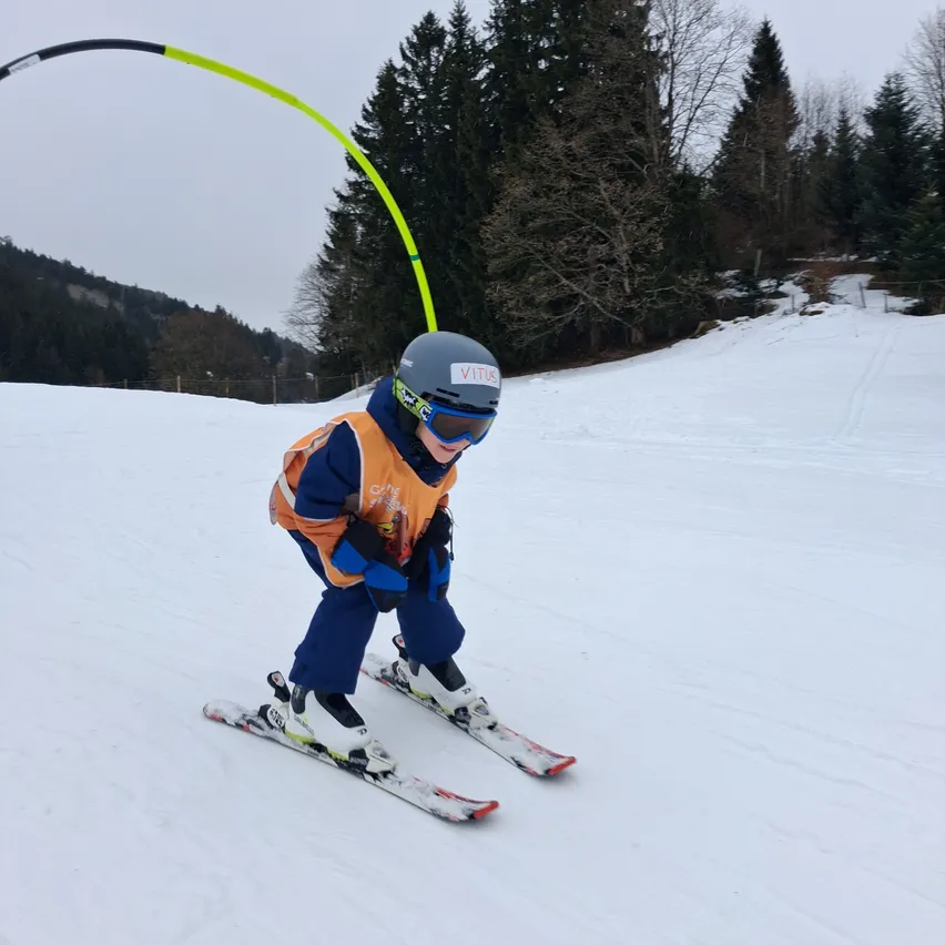 Ein junger Skifahrer navigiert eine Slalomstrecke auf einer verschneiten Piste, trägt einen Helm, eine Sonnenbrille und ein leuchtendes orangefarbenes und blaues Outfit.
