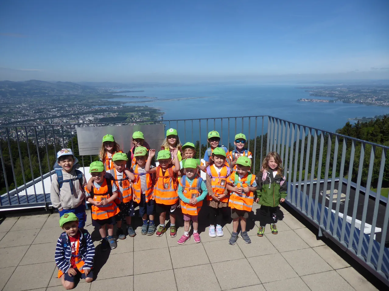 Eine Gruppe von Kindern in Warnwesten posiert auf einem Balkon mit Blick auf eine Stadt und das Meer.