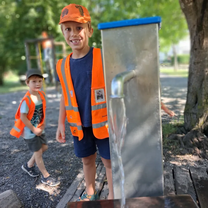 Zwei junge Jungen, beide mit orangefarbenen Westen, stehen neben einem Wasserbrunnen in einem Park und lächeln in die Kamera.