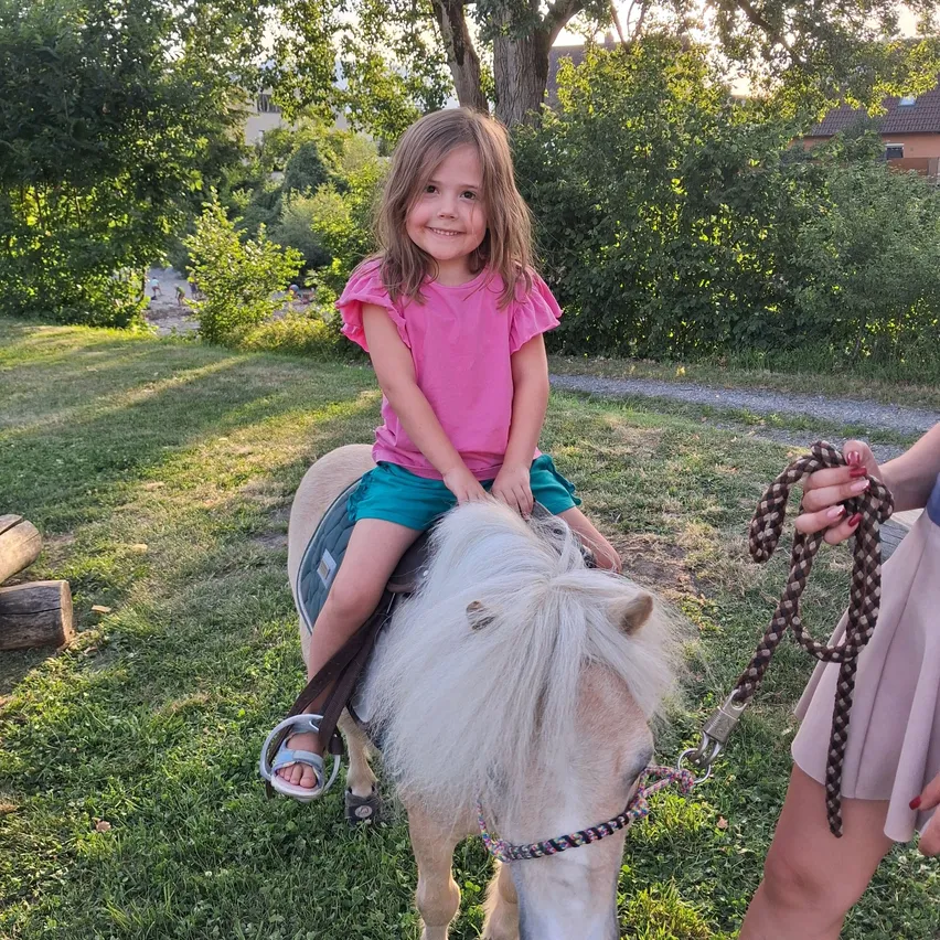 Ein kleines Mädchen reitet auf einem weißen Pony im Park, gehalten von einem Erwachsenen, der eine Leine hält. Das Pony hat ein farbenfrohes Zaumzeug. Bäume und ein Weg sind im Hintergrund zu sehen.