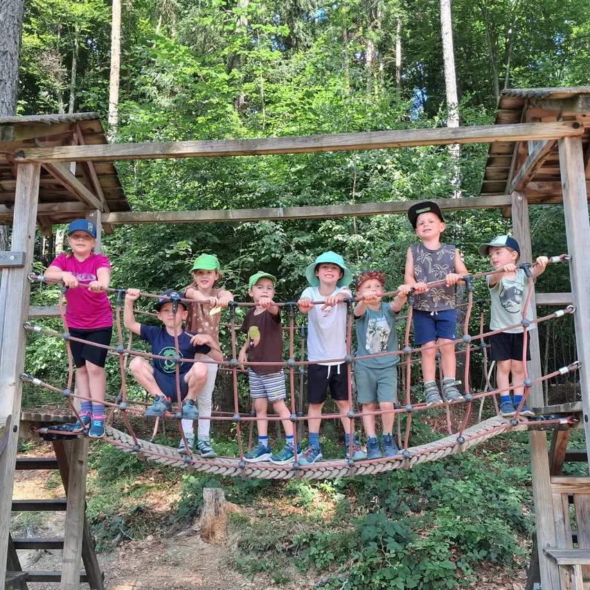 Eine Gruppe von Kindern steht auf einer Holzbrücke im Wald und hält sich an den Seilen fest. Sie lächeln alle und scheinen den sonnigen Tag zu genießen.