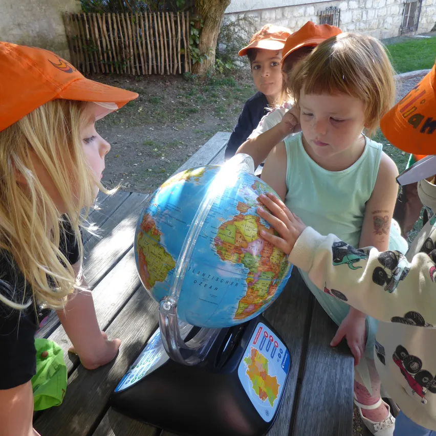 Eine Gruppe junger Kinder, die orangefarbene Mützen tragen, sind um einen Globus versammelt, wobei ein Kind es hält und ein anderes auf einen Ort zeigt.