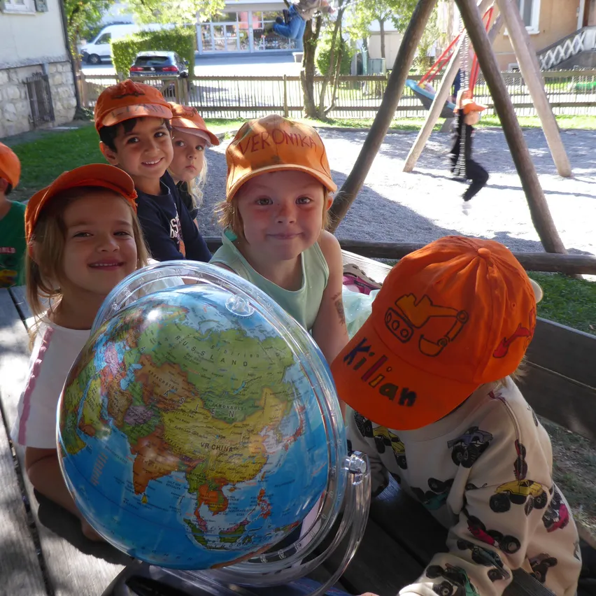 Fünf Kinder, alle mit orangefarbenen Mützen, sitzen auf einer Bank mit einem Globus im Vordergrund. Dahinter ist ein Spielplatz mit Schaukeln und einer Rutsche zu sehen.