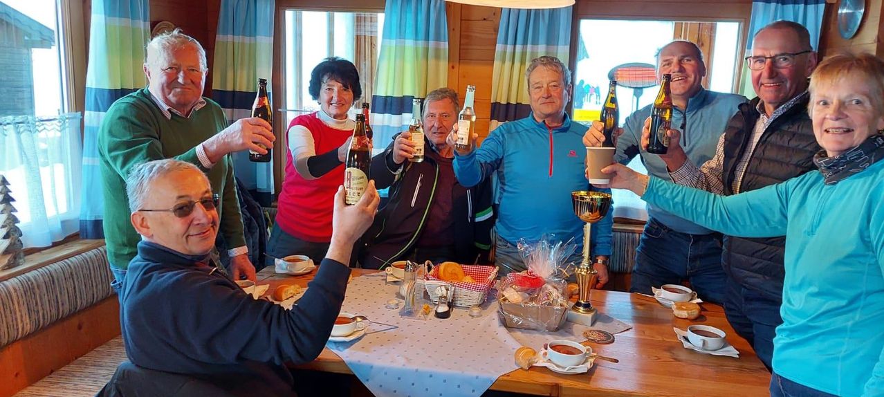 Several adults, smiling, are toasting with beer bottles in a dining room with a table covered with bread, pastries, and cups. A trophy is present on the table.