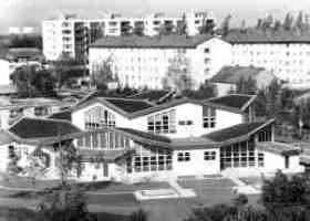 Ein schwarz-weißes Luftbild eines modernen Gebäudes mit vielen Fenstern und einem schrägen Dach. Bäume umgeben es, und andere Gebäude sind im Hintergrund zu sehen.
