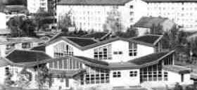 Ein schwarz-weißes Luftbild eines modernen Gebäudes mit vielen Fenstern und einem schrägen Dach. Bäume umgeben es, und andere Gebäude sind im Hintergrund zu sehen.