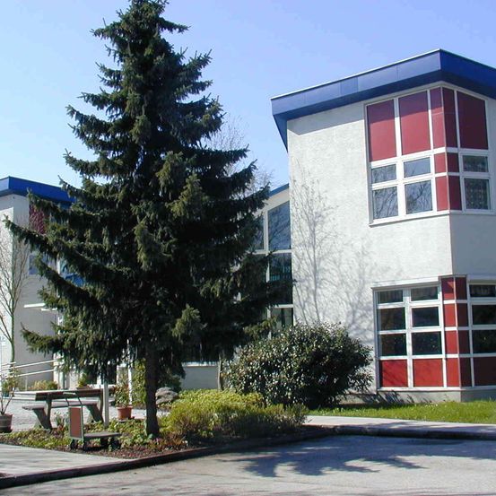 Ein modernes Gebäude mit weißen Wänden und roten Akzenten. Ein hoher Baum steht im Vordergrund mit einer Bank und Topfpflanzen.