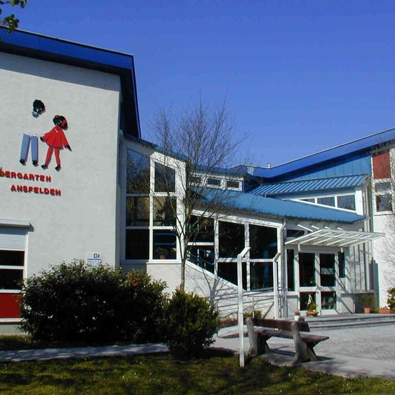 Ein Gebäude mit einer Wandmalerei von zwei Kindern und dem Text 'Kindergarten Ansfelden'. Es hat Glasfenster und ein blaues Dach. Ein Bank vorn.