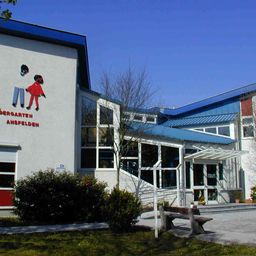 Ein Gebäude mit einer Wandmalerei von zwei Kindern und dem Text 'Kindergarten Ansfelden'. Es hat Glasfenster und ein blaues Dach. Ein Bank vorn.