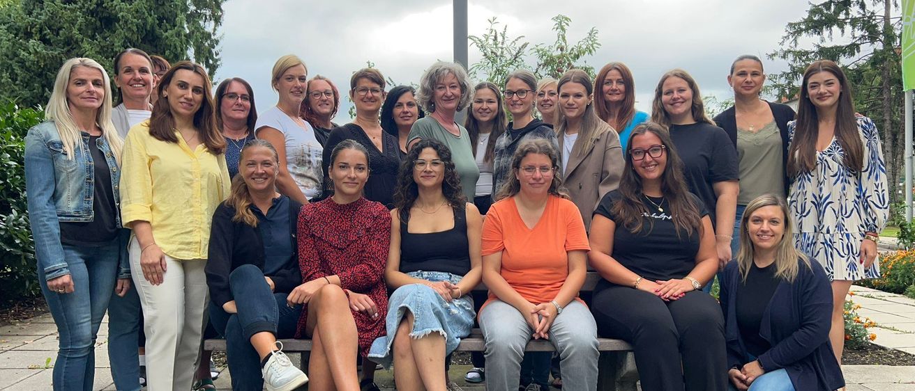 Eine Gruppe von Frauen in Freizeitkleidung posiert für ein Foto im Freien, mit einem Steinweg und Bäumen im Hintergrund.