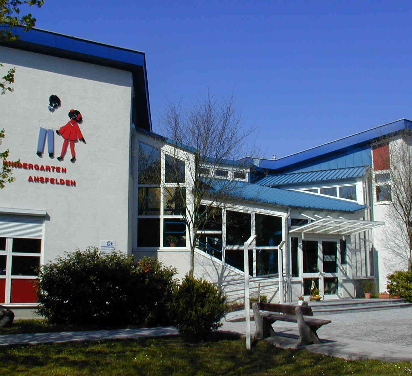 Der Eingang eines Kindergartengebäudes mit einer Wandmalerei von zwei Kindern. Das Gebäude hat ein blaues Dach und weiße Wände. Ein Bank ist davor platziert.