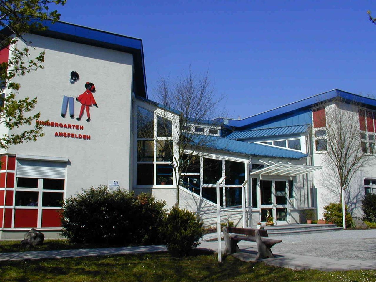 Der Eingang eines Kindergartengebäudes mit einer Wandmalerei von zwei Kindern. Das Gebäude hat ein blaues Dach und weiße Wände. Ein Bank ist davor platziert.