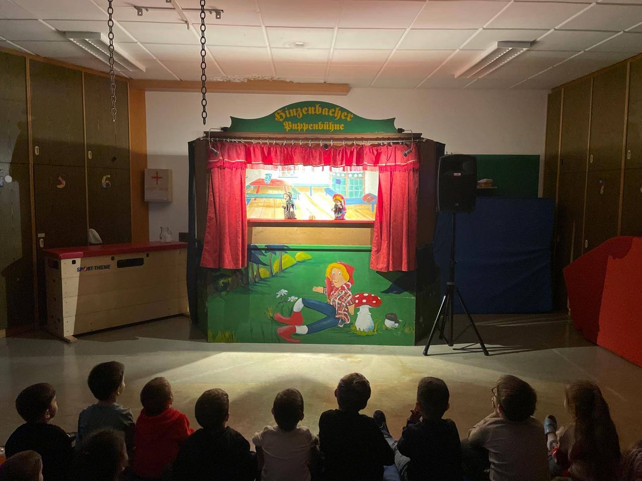 Kinder schauen sich ein Puppentheater mit einem farbenfroh bemalten Hintergrund auf der Bühne an, das eine Figur und Pilze zeigt.