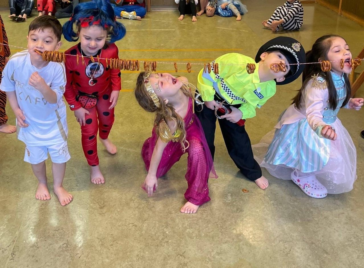Kinder in Kostümen, darunter eine Marienkäferin und ein Polizeibeamter, spielen mit einer Pretzel-Schnur in einer Halle. Einige sitzen und stehen im Hintergrund.