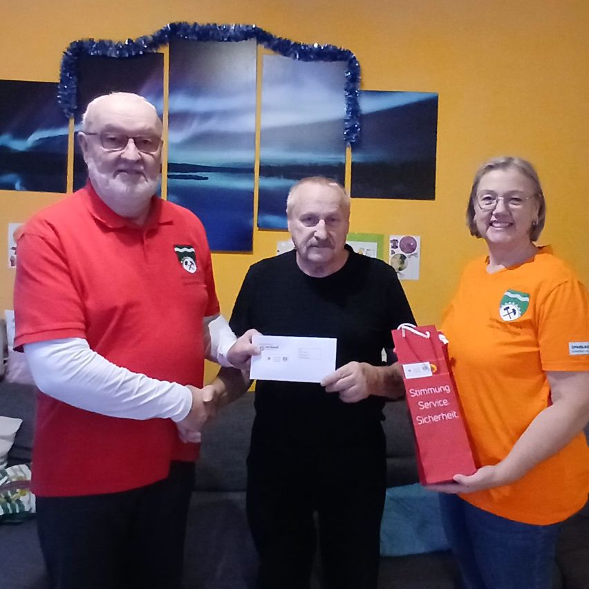 Drei Personen in einem Raum, zwei in orangefarbenen Shirts, die einen roten Beutel halten, einer in einem schwarzen Shirt, der einen Scheck hält.