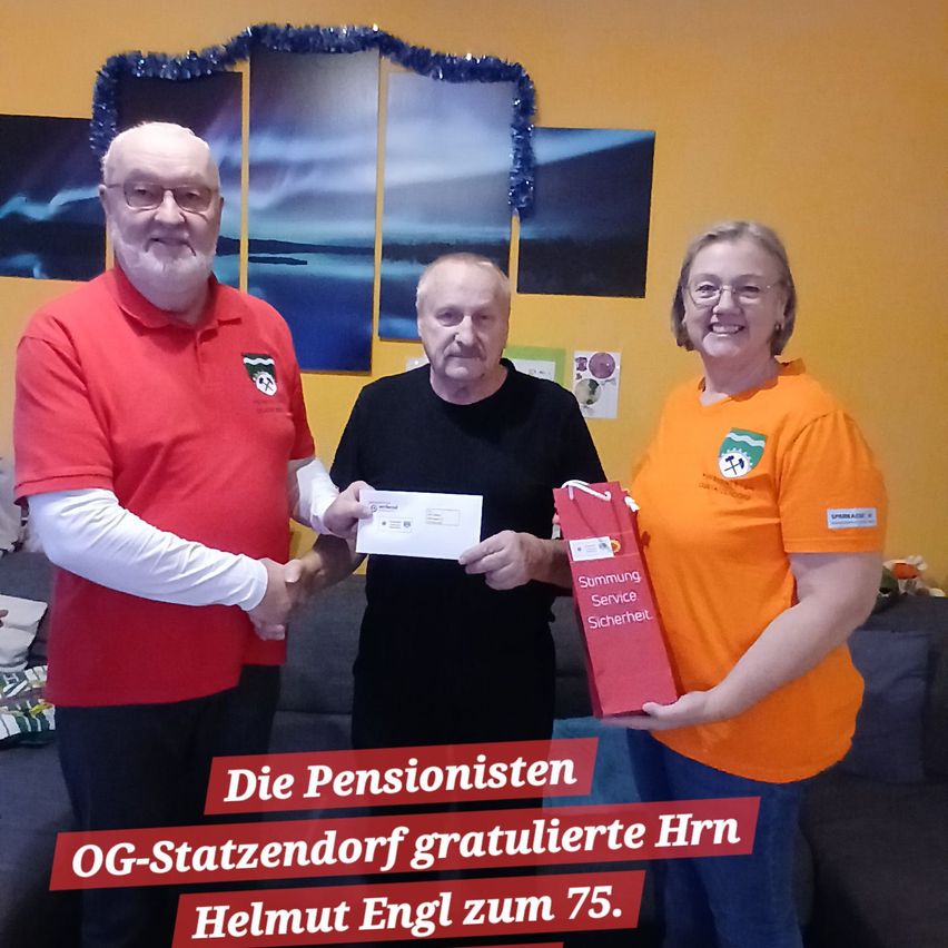 Drei Menschen stehen eng beieinander und lächeln. Ein Mann in einem roten Shirt hält einen Scheck, ein anderer Mann in einem schwarzen Shirt hält einen roten Geschenkbeutel und eine Frau in einem orangefarbenen Shirt lächelt. Hinter ihnen hängen gerahmte Bilder an einer gelben Wand.