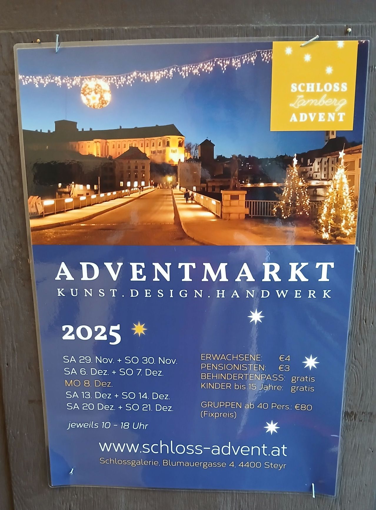 Plakat für den Adventmarkt 2025 im Schloss Lamberg, zeigt eine geschmückte Straße mit Gebäuden, Bäumen und Lichtern. Eintrittspreise für Erwachsene, Senioren und Kinder aufgeführt.