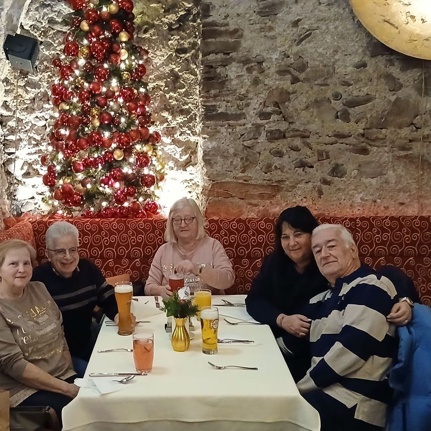 Fünf Personen sitzen um einen Tisch mit Getränken, einer Blumenvase und Besteck. Dahinter steht ein roter Weihnachtsbaum.