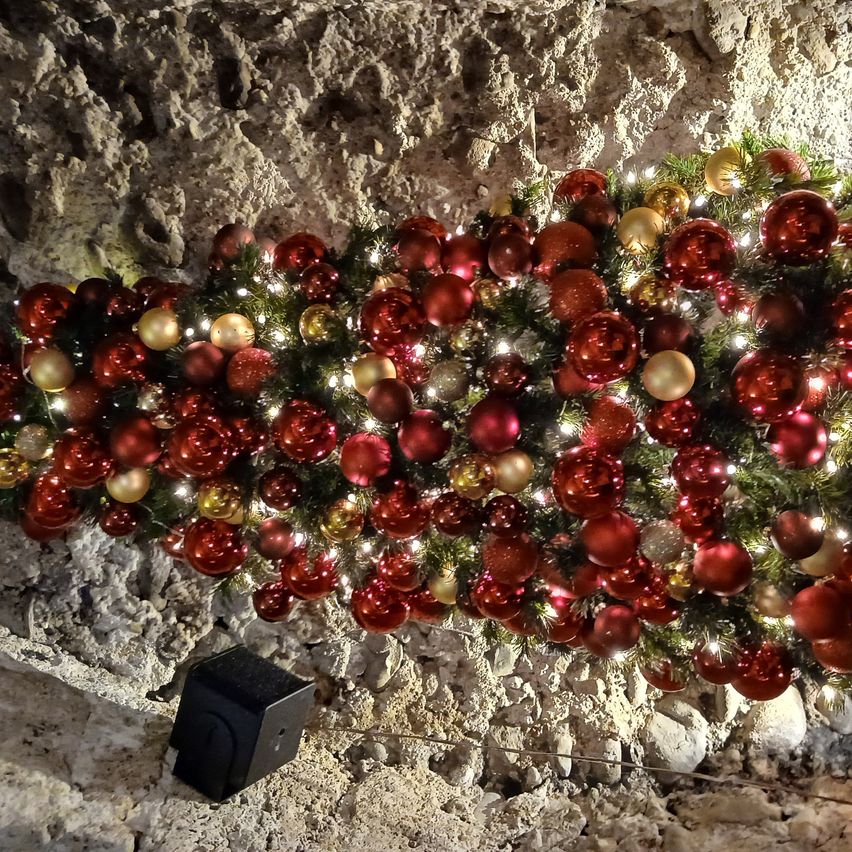 Ein Weihnachtsbaum aus roten und goldenen Kugeln ist an einer felsigen Wand angebracht. Der Baum ist mit Lichtern und kleinen grünen Blättern geschmückt. Unter dem Baum ist ein schwarzer Lautsprecher zu sehen.