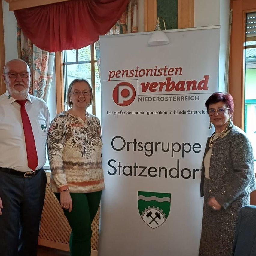 Drei Personen stehen neben einem Banner mit der Aufschrift 'pensionisten verband'. Sie alle lächeln und scheinen sich bei einer Veranstaltung zu befinden.