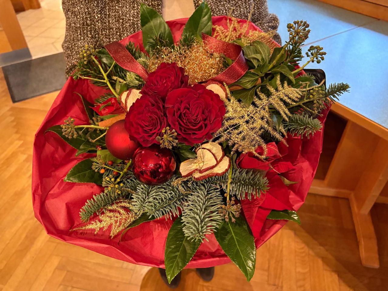 Ein roter Blumenstrauß mit mehreren Rosen, grünen Blättern und goldenen Akzenten, geschmückt mit roten Ornamenten und einem roten Band. Er ruht auf einer Holzoberfläche.