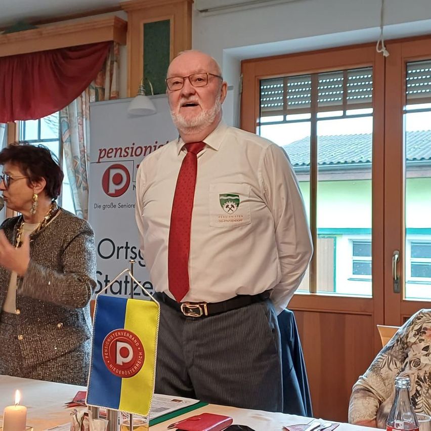 Ein älterer Mann steht mit einer Flagge und einer Kerze auf einem Tisch, während eine Frau in ein Mikrofon spricht. Dahinter steht ein Schild mit dem Wort 'pension' und einem Logo. Im Hintergrund befinden sich Fenster und eine Tür.