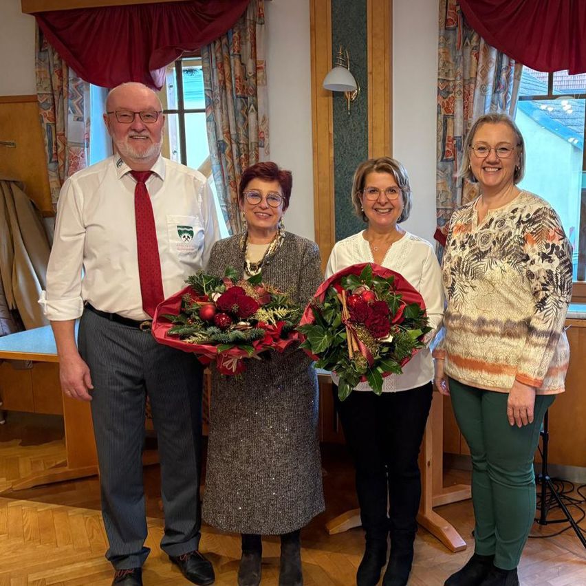 Vier Menschen, zwei Männer und zwei Frauen, stehen zusammen in einem Raum. Sie halten Blumensträuße und scheinen für ein Foto zu posieren. Der Raum hat Holzböden und Wände, die mit Vorhängen geschmückt sind.