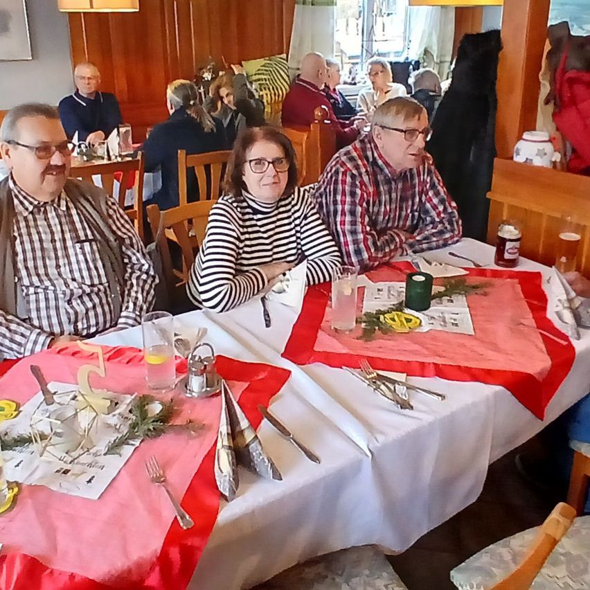 Drei Menschen sitzen in einem Restaurant an einem Tisch, warm gekleidet. Ein Mann links, eine Frau in der Mitte und ein Mann rechts. Der Tisch ist mit Besteck, Servietten und Getränken gedeckt.