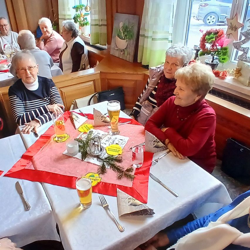 Mehrere ältere Frauen sitzen um einen Tisch in einem Restaurant, lächeln und unterhalten sich. Der Tisch hat Biergläser, eine Serviette und einen roten Stoff. Das Restaurant hat Holzwände und ein Fenster mit Blick auf ein Auto.