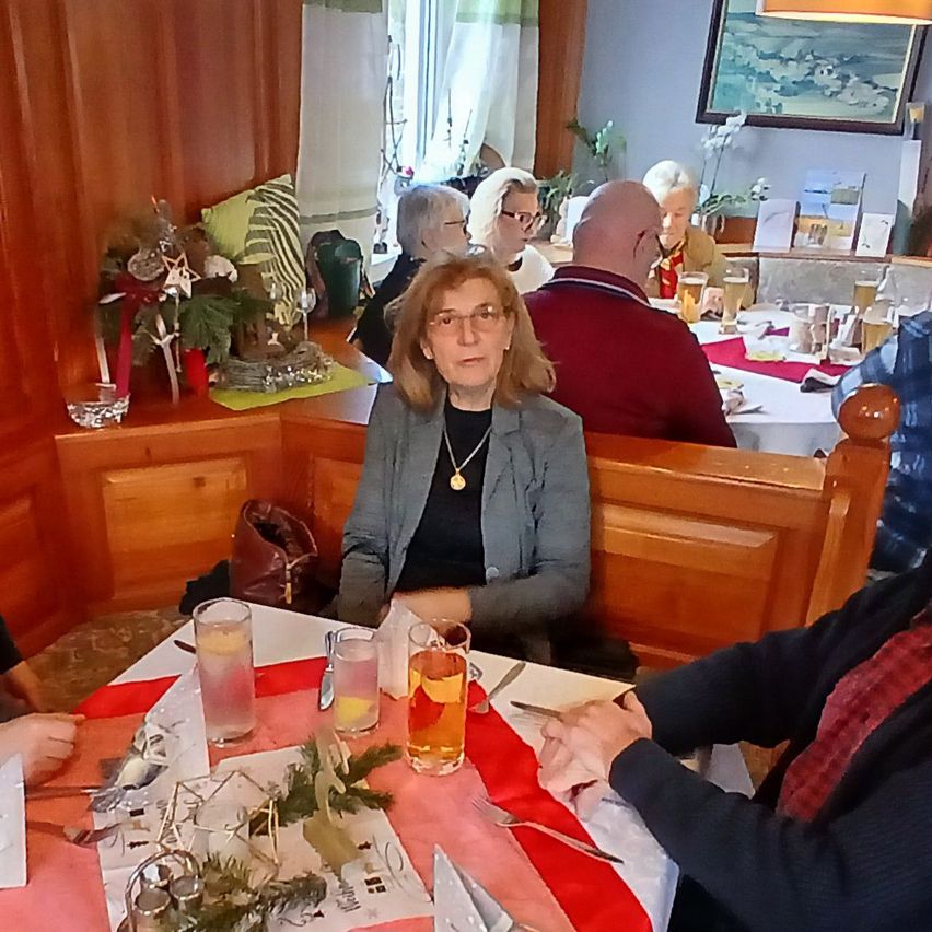 Eine Gruppe von Menschen sitzt an einem Tisch in einem Restaurant. Eine Frau mit Brille ist im Vordergrund. Hinter ihr sitzt ein Mann mit kahlem Kopf. Eine Topfpflanze steht auf dem Regal hinter ihnen.