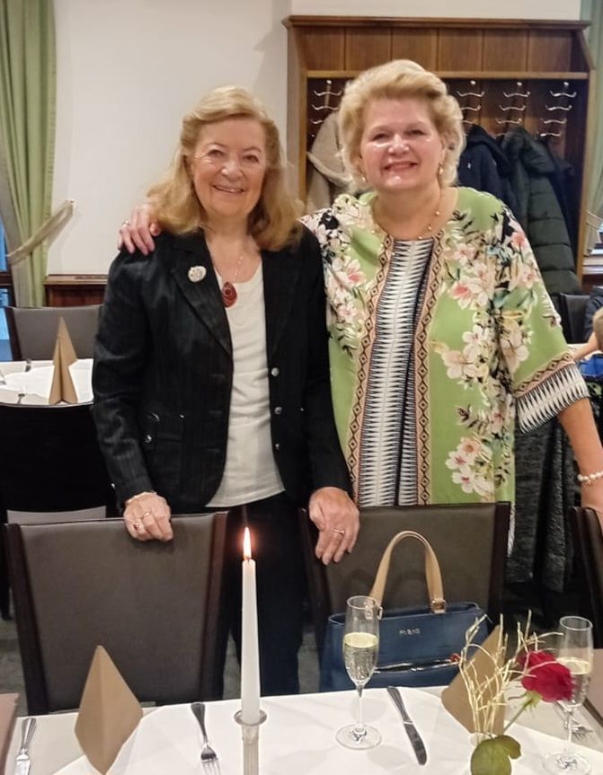 Zwei Frauen lächeln, eine in einer schwarzen Jacke und die andere in einem grünen Blumentopf, stehen neben einem Tisch mit einer brennenden Kerze, Weingläsern und einer blauen Handtasche.