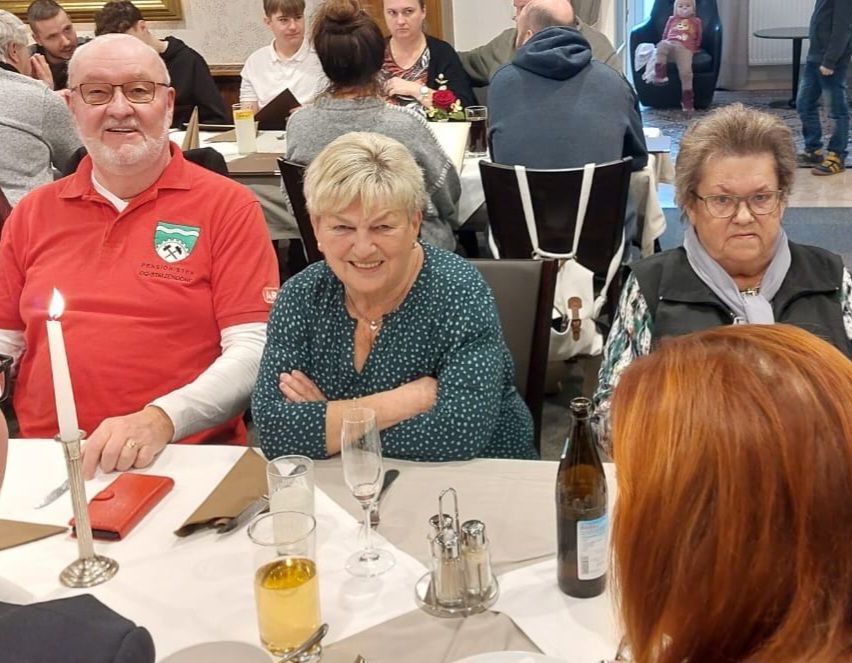 Eine Gruppe von Menschen versammelt sich in einem Restaurant. Ein Mann in einem roten Hemd sitzt mit einer Frau in einem gepunkteten Oberteil. Hinter ihnen schaut eine Frau mit Brille und Schal zu. Ein Baby sitzt in einem Kinderwagen in der Nähe der Wand.