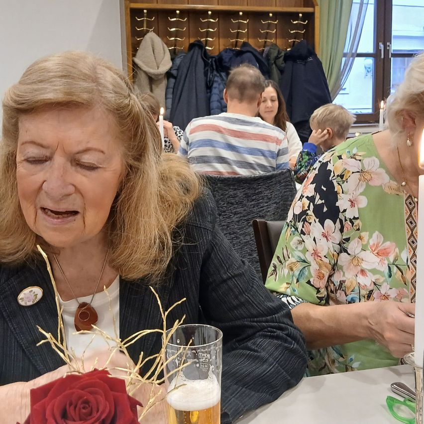 Eine ältere Frau sitzt an einem Tisch mit einer Rose und einem Glas Bier, scheinbar emotional. Ein Mann in einem gestreiften Shirt und eine Frau in einem Blumenkleid sitzen hinter ihr. Ein Kind ist ebenfalls in der Nähe sitzend und Mäntel hängen an einer Garderobe.