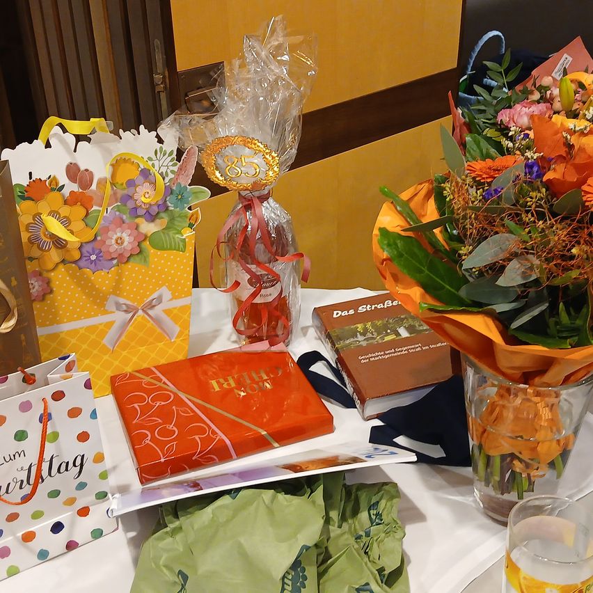 Ein Tisch mit mehreren Geschenken und Gegenständen. Ein Blumenstrauß ist in einer Glasvase. Ein Buch, eine Geschenktüte und eine verpackte Flasche sind ebenfalls auf dem Tisch.