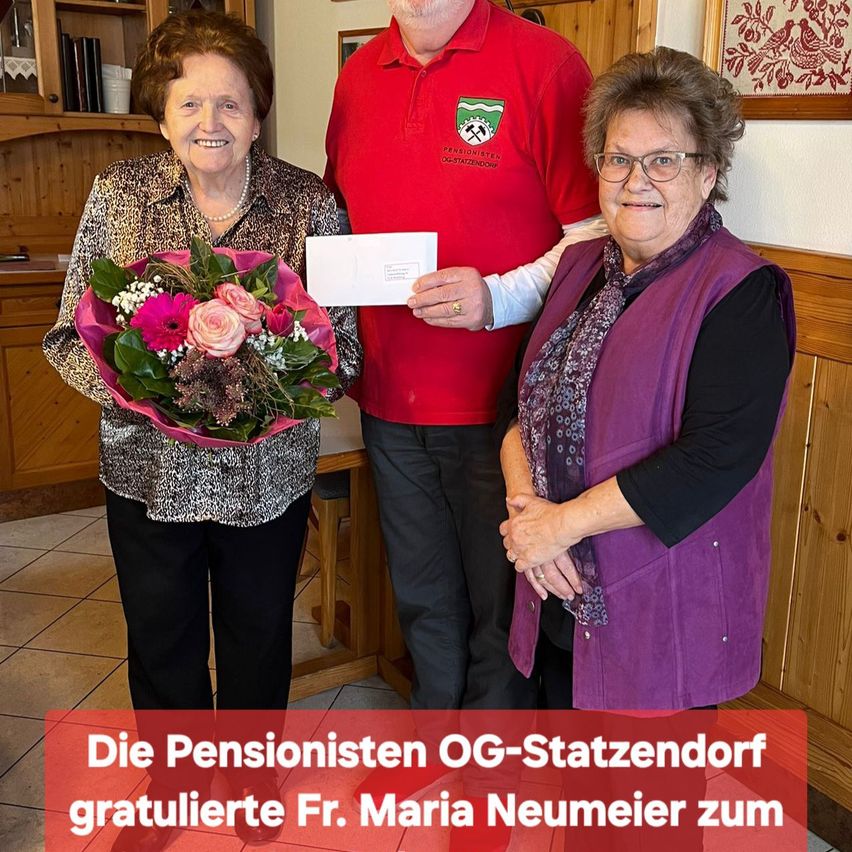 Eine Frau hält einen Blumenstrauß und steht mit zwei anderen. Der Mann hält ein Papier und sie stehen in einem Raum mit einer Holzwand und einem Regal.